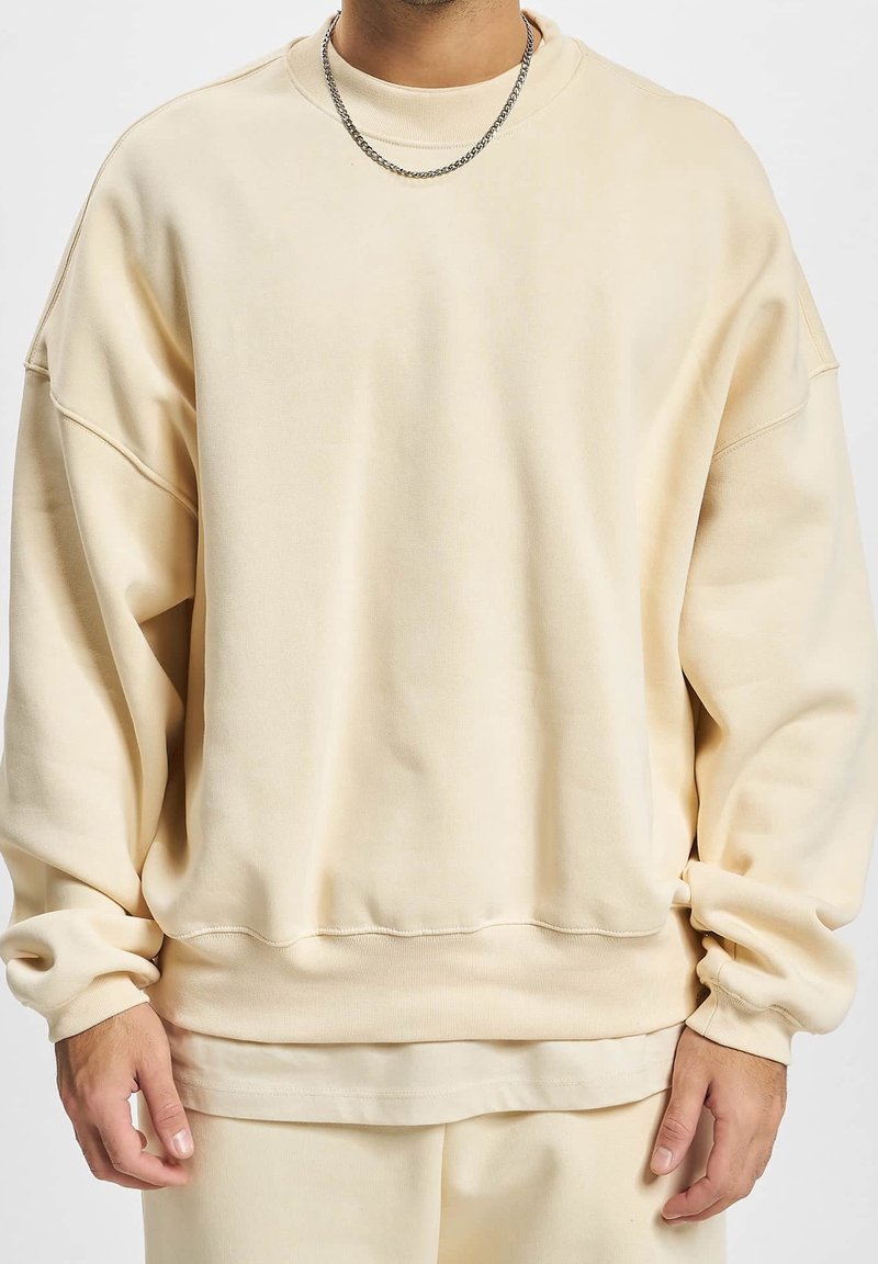 Felpa beige oversize con spalle scese, colletto a costine e maniche lunghe con polsini. Tessuto morbido con una texture liscia.