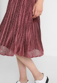 Plisserad midikjol i glänsande djupröd tyg. Genomskinlig textur med skimrande effekt, kombinerad med vita skor med svarta detaljer.