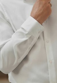 Weißes Hemd aus strukturiertem Stoff, mit klassischer Knopfleiste und umgeschlagenem Ärmel mit einem einzelnen Knopf zum Schließen.