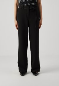 Kobieta ubrana w czarne szerokie spodnie z wysokim stanem, z czarnym paskiem ozdobionym kwiatem, w połączeniu z czarnymi czółenkami.