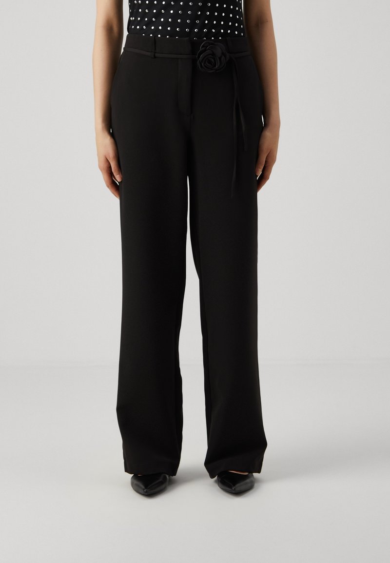 Kobieta ubrana w czarne szerokie spodnie z wysokim stanem, z czarnym paskiem ozdobionym kwiatem, w połączeniu z czarnymi czółenkami.