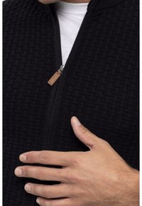 Schwarze Zip-Up-Jacke aus strukturiertem Stoff, mit Stehkragen und einem Leder-Zipper. Eine Hand liegt auf der Jacke und hebt das Design hervor.