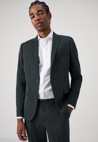 Dark green suit jacket with a notched lapel, single button front, and a chest pocket. Worn over a white button-up shirt.