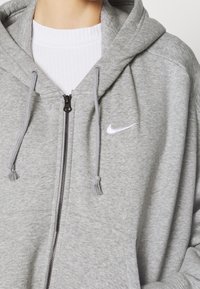 Sweat à capuche gris zippé en matière douce. Il présente une fermeture éclair à l'avant, une capuche avec cordon de serrage, et un logo Nike blanc sur la poitrine.