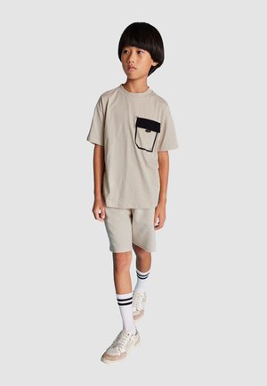 T-shirt beige con tasca nera, abbinato a pantaloni corti beige. Il modello indossa anche calze bianche con strisce nere e sneakers chiare.