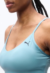 Γυναίκα που φοράει ανοιχτό μπλε αθλητικό τοπ Puma με λεπτές τιράντες και πλεγμένα μαλλιά, εμφανιζόμενη από τον λαιμό έως το στήθος σε λευκό φόντο.