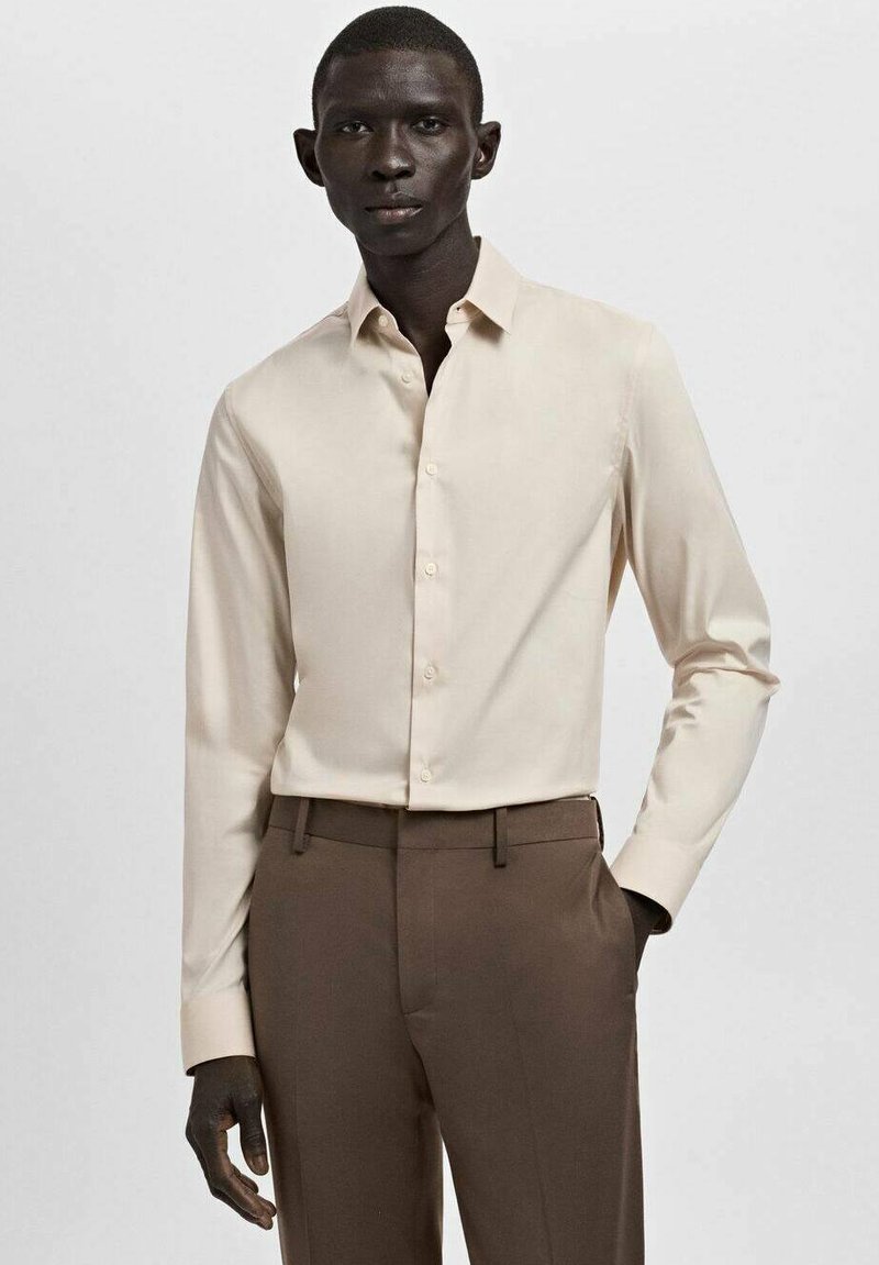 Chemise beige à manches longues avec un col pointu, une fermeture à boutons sur le devant et une texture lisse, associée à un pantalon marron sur mesure.