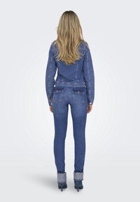 Kvinna med långt hår vänd bort, iförd en blå jeansjacka och åtsittande jeans, som står framför en ljus bakgrund och visar upp sin outfit.