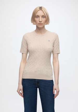 Donna bionda in piedi di fronte con indosso un top beige a maniche corte lavorato a maglia e jeans blu scuro su uno sfondo chiaro e semplice.