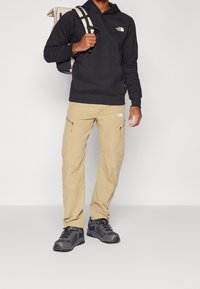 Sudadera negra con capucha, pantalones cargo color beige con bolsillos con cremallera, zapatos de senderismo negros con suelas texturizadas y una mochila beige con correas.