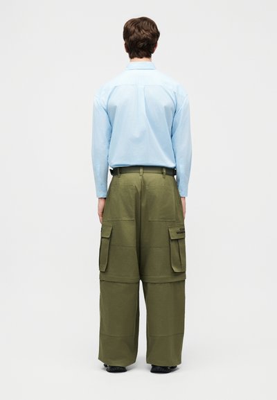 Άνδρας με πλάτη γυρισμένη, φοράει ανοιχτό μπλε πουκάμισο με μακριά μανίκια και φαρδιά χακί παντελόνια τύπου cargo με πολλές τσέπες, στέκεται μπροστά σε λευκό φόντο.