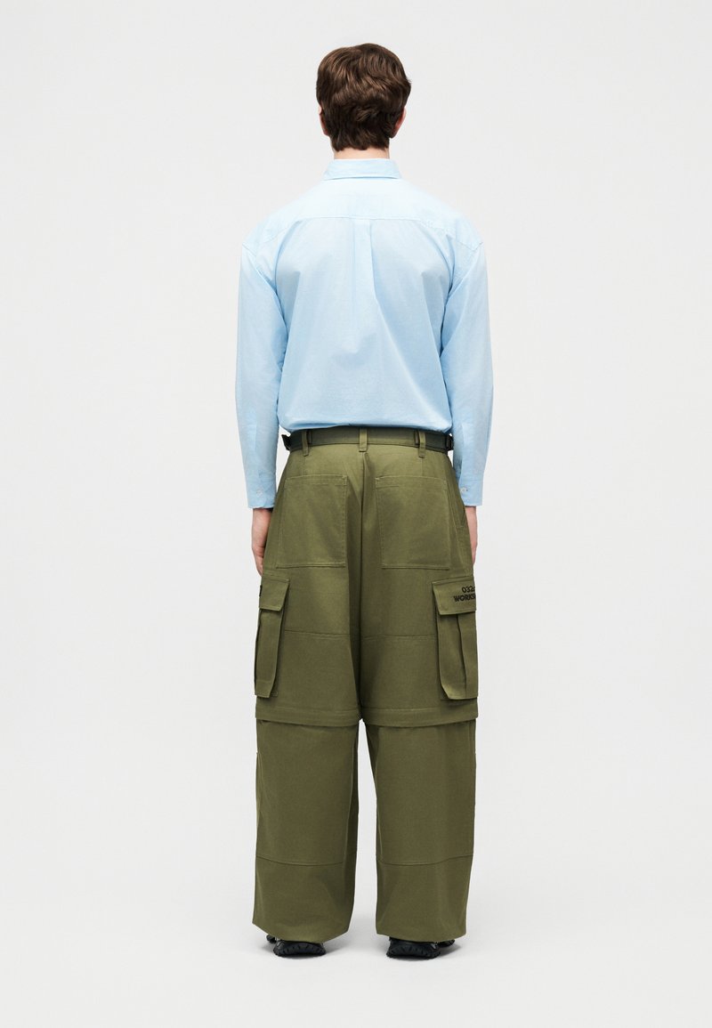 Hombre de espaldas vistiendo una camisa de manga larga azul claro y pantalones cargo verdes oliva de gran tamaño con múltiples bolsillos, de pie sobre un fondo blanco.