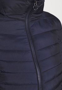 Giacca puffer blu navy con colletto alto, trapuntatura verticale e zip frontale con un tab circolare, realizzata in tessuto liscio e lucido.