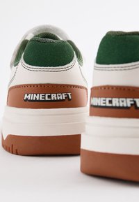 Baskets blanches et marron avec des accents verts ; semelle en caoutchouc texturée ; logo "MINECRAFT" sur le talon ; matériaux lisses et en daim utilisés.