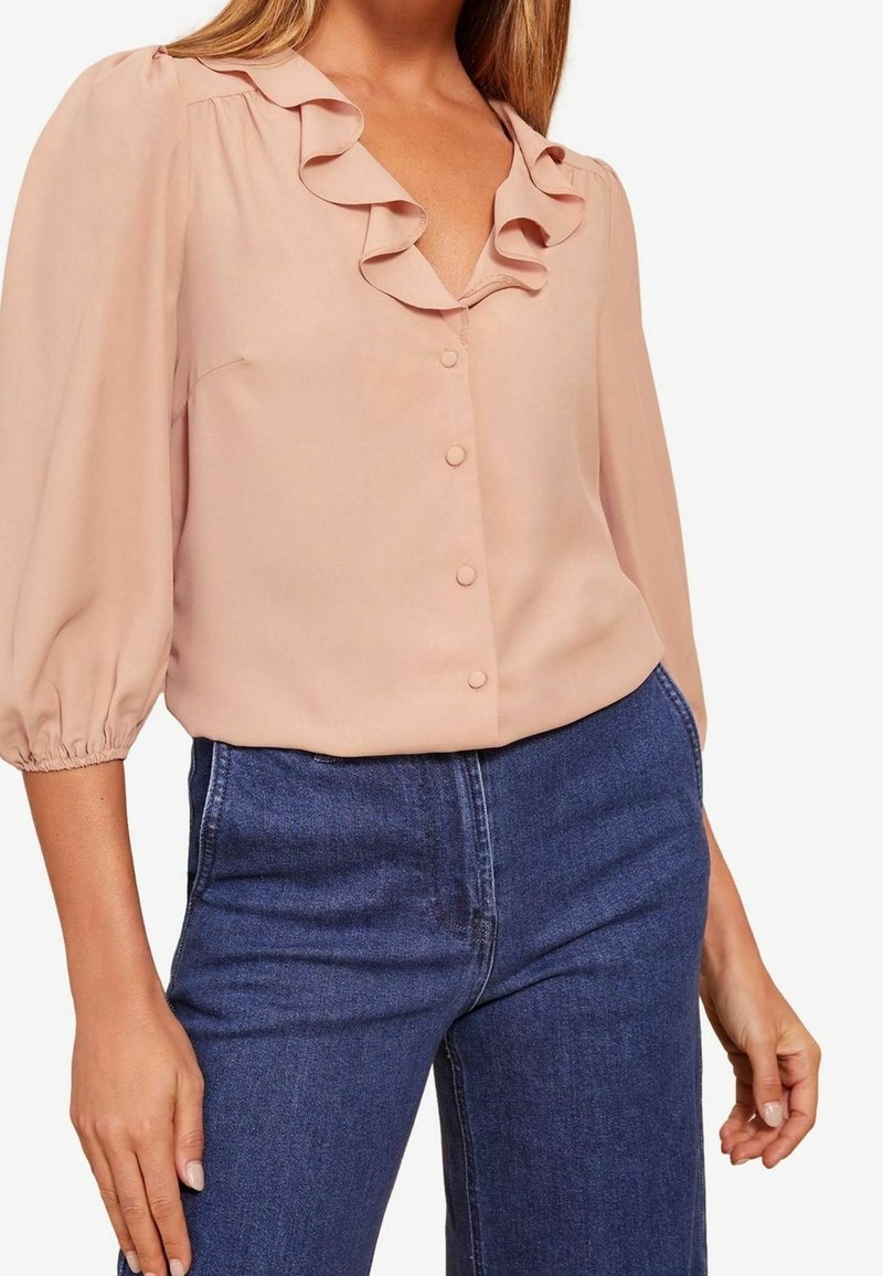 Blouse rose clair avec un col en V à volants et des boutons à l'avant, manches bouffantes. Portée avec un jean taille haute bleu foncé.