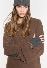 Barna, texturált, túlságosan nagy pulóver hosszú ujjal. Szürke ujj nélküli kesztyűk hangsúlyozzák a mandzsettákat. A modellen egy sötétszürke kötött sapka van.