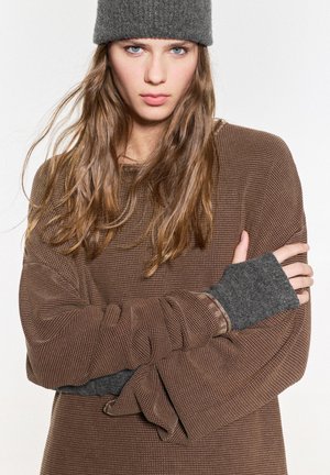 Brązowy, teksturowany, oversize sweter z długimi rękawami. Szare rękawiczki bez palców podkreślają mankiety. Model ma na sobie ciemnoszary, dziany beanie.