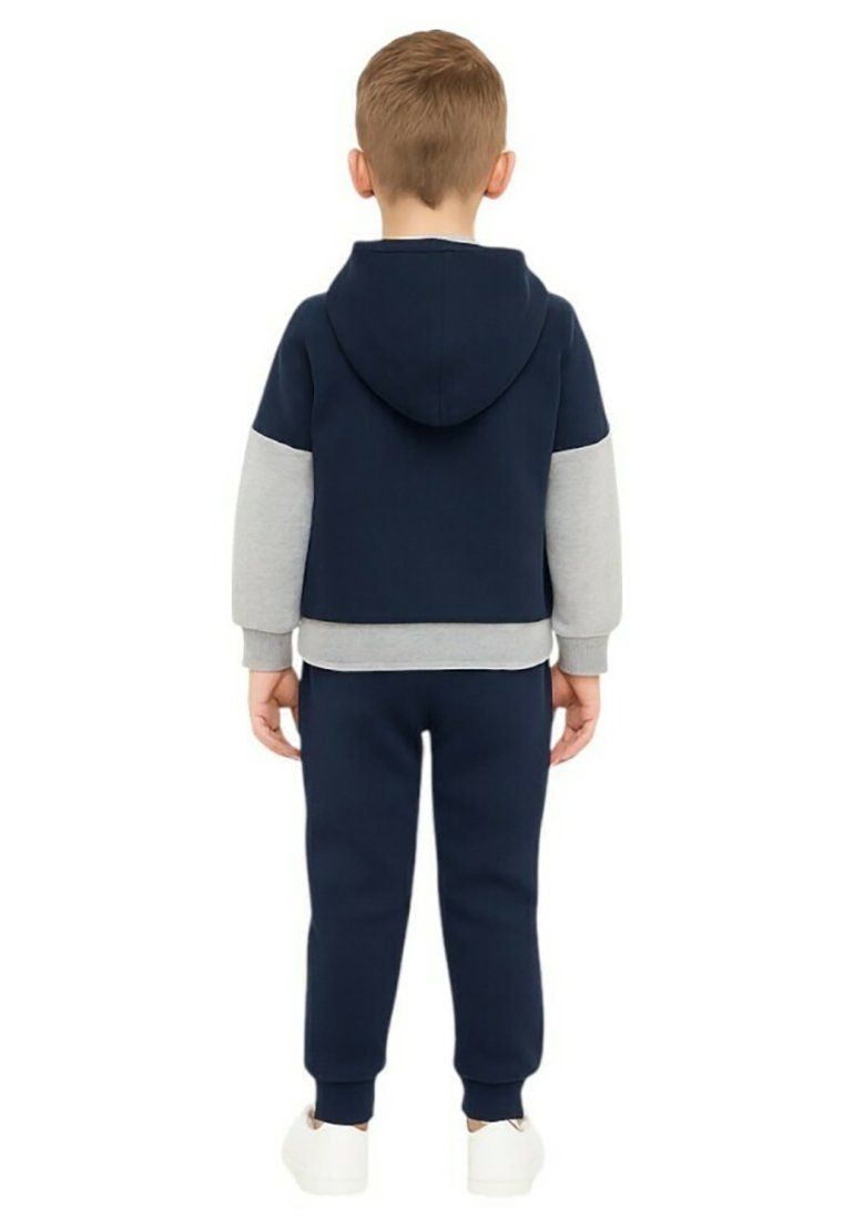 Enfant debout de dos, portant un sweat à capuche bleu marine et gris avec un pantalon de survêtement bleu marine assorti et des baskets blanches.