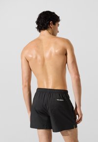 Čierne plavecké šortky s elastickým pásom a logom na zadnej strane, vyrobené z ľahkej látky, prezentované na nahom tele na neutrálnom pozadí.