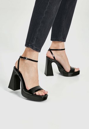Czarne sandały na wysokim obcasie z błyszczącym wykończeniem, szerokim paskiem z przodu, paskiem wokół kostki oraz masywnym słupkowym obcasem. Otwarty przód z platformą.