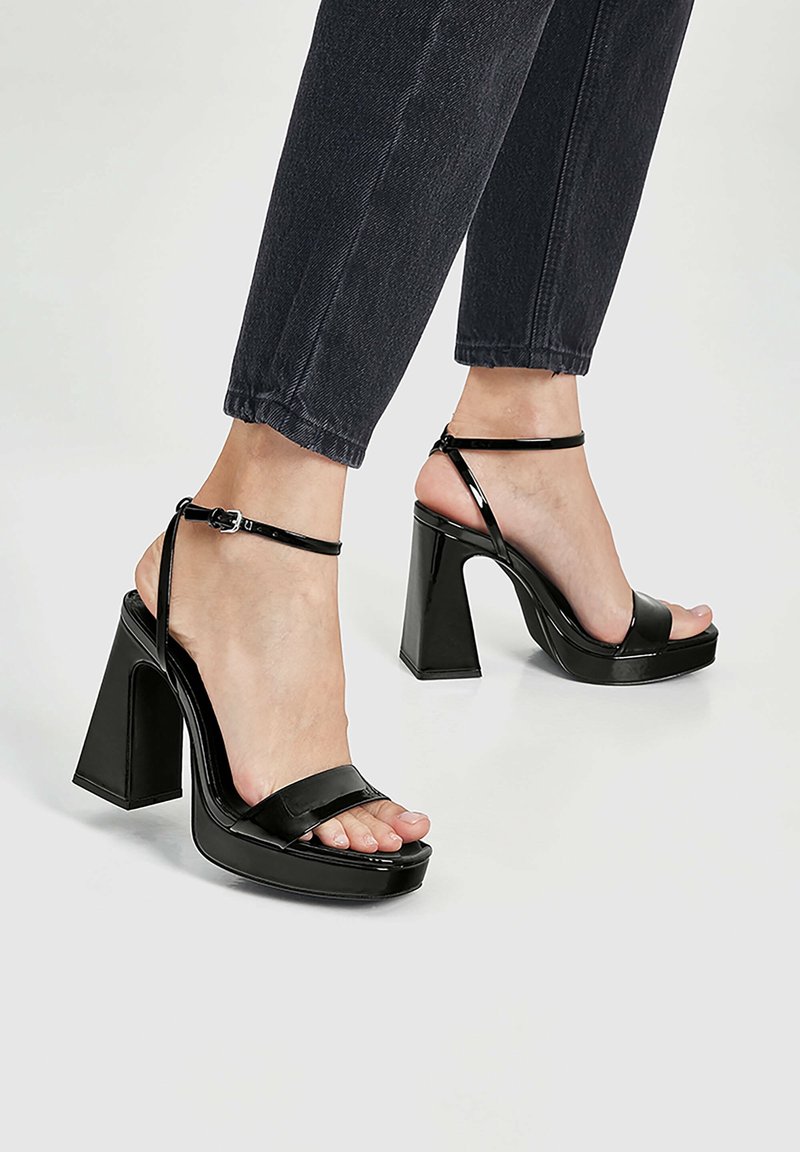 Sandales à talons hauts noires avec une finition brillante, large bride avant, bride à la cheville et talon épais. Bout ouvert avec une semelle compensée.