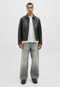 Black leather jacket with two front pockets, worn over a white polo shirt. Loose-fitting faded blue jeans and brown boots complete the look.