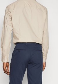 Beige Langarmhemd mit Knopfmanschetten, kombiniert mit marineblauen Hosen mit zwei Gesäßtaschen und Knopfverschlüssen. Glatte Stofftextur.