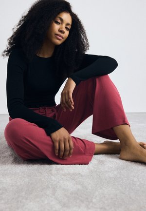 Zwarte langemouwen top gedragen met losse, ribgebreide, bordeauxrode broek. Onderwerp zit op een lichtgrijze vloerbedekking, op blote voeten, met krullend haar.