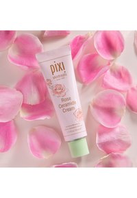 Pixi Rose Ceramide Cream-tube, rosa med roseillustrasjoner, grønn kork, omgitt av rosa rosenblad på en lys bakgrunn.
