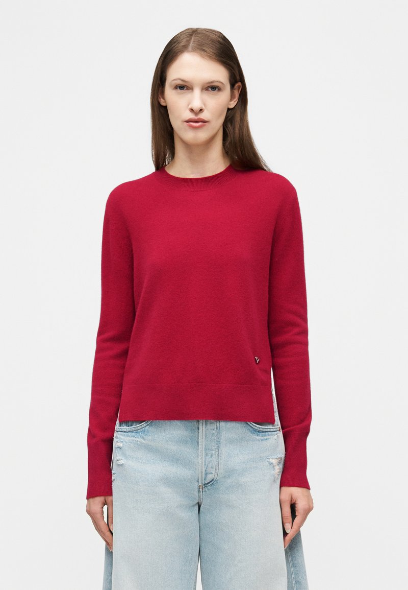 Γυναίκα με ίσια καστανά μαλλιά που φοράει κόκκινο πουλόβερ με μακριά μανίκια και ανοιχτό μπλε σκισμένο τζιν, στέκεται μπροστά σε απλό λευκό φόντο.