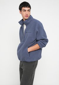 Bluza polarowa w miękkim niebieskim kolorze z kołnierzem na pół suwaka, przednią kieszenią na zamek oraz kieszeniami bocznymi, z podwiniętymi rękawami i luźnym krojem.