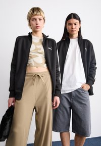 Et par personer iført matchende sorte zip-up jakker med hvide detaljer. Den ene har en beige mønstret cropped top; den anden en ensfarvet hvid t-shirt.