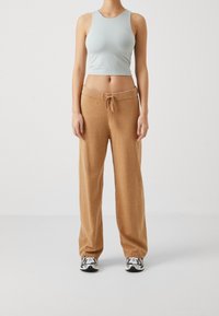 Woman wearing beige drawstring pants, light blue sleeveless crop top, and white sneakers standing against plain white background.