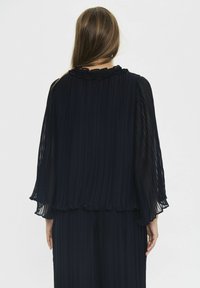 Donna con capelli lunghi che indossa una blusa plissettata blu navy con colletto arricciato e maniche larghe, vista da dietro contro uno sfondo semplice.