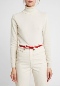 Maglione a collo alto color crema in morbido tessuto a maglia, abbinato a jeans bianchi a vita alta e una cintura in pelle rossa con dettaglio a fiocco.