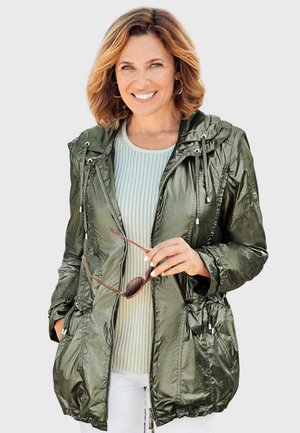 Leichte olivgrüne Jacke mit glänzender Oberfläche, Kordelzugkapuze und gerafftem Bund. Über einem hellgrünen, gerippten Oberteil getragen.
