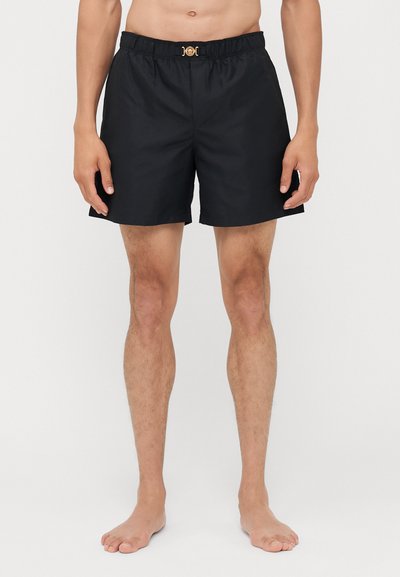 Shorts de bain noirs avec une taille élastique et des détails en métal doré à l'avant. Texture lisse, longueur au-dessus du genou. Design simple.