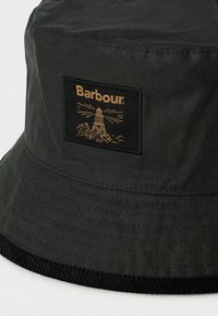 Tmavě zelený klobouk s širokým okrajem a černým lemem, s obdélníkovou nášivkou zobrazující maják a slovo "Barbour" ve zlaté barvě.