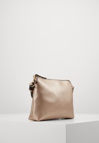 Piccolo borsa a tracolla in pelle metallizzata rose gold, con texture, chiusura a zip e tracolla nera regolabile, esposta su un piedistallo bianco.