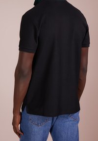 Mężczyzna ubrany w czarną koszulkę polo z krótkim rękawem oraz niebieskie dżinsy, pokazany od tyłu na jednolitym tle.