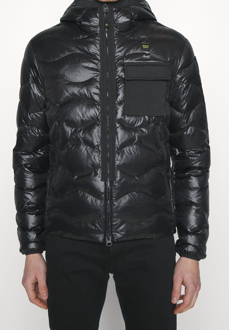 Veste noire matelassée avec une finition brillante, capuche, fermeture éclair et poche poitrine. Le design présente des motifs matelassés incurvés et des poignets élastiques.