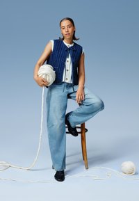 Gilet en tricot bleu foncé avec des côtes verticales, boutons blancs, associé à un pantalon en jean clair à jambes larges, tenant de la laine blanche. Chaise en bois simple à l'arrière-plan.