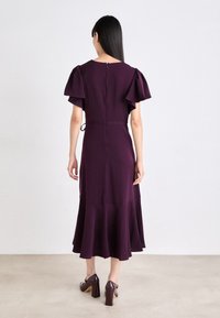 Rochie midi purpurie cu mâneci bufante, bust ajustat, tiv evazat și fermoar la spate. Asortată cu pantofi cu toc maro, cu o textură netedă.