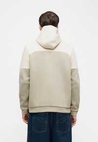 Άνδρας που φοράει φούτερ με κουκούλα διχρωμία σε μπεζ αποχρώσεις, με την κουκούλα ανασηκωμένη, συνδυασμένο με σκούρο μπλε τζιν, από πίσω σε απλό φόντο.