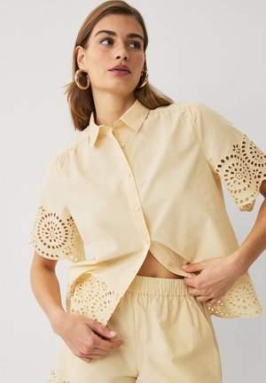 Kvinde iført beige kortærmet skjorte med øjekutter-udskæringer og matchende elastiske bukser justerer skjortens kant.