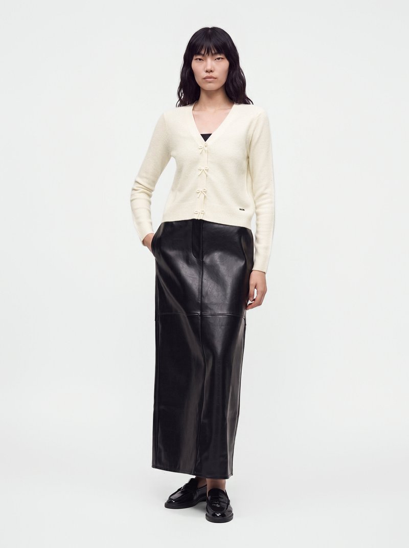 Femme portant un cardigan crème boutonné et une longue jupe en cuir noire, debout les mains dans les poches.