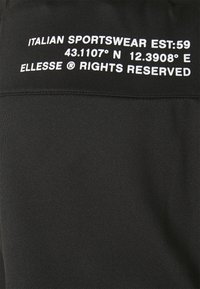 Svart sportklädesmaterial med vit text som säger "ITALIAN SPORTSWEAR EST:59 43.1107° N 12.3908° E ELLLESSE ® RÄTTIGHETER FÖRBEHÅLLNA."