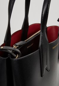 Svart läder tote-väska med texturerad yta, röd inredning, guldfärgad dragkedjestängning och präglad logotyp. Två robusta handtag för bärande.