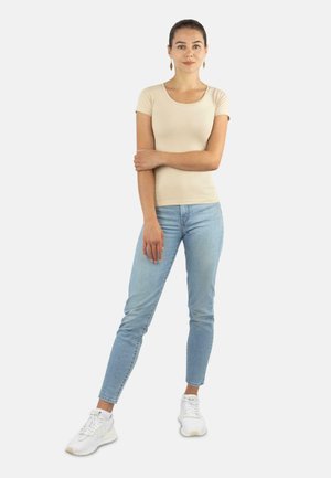 Frau steht mit einem Arm quer über dem Oberkörper, trägt ein beige anliegendes T-Shirt, hellblaue Jeans, weiße Turnschuhe und goldene Ohrringe.