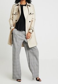 Beżowy płaszcz trencz z czarnymi guzikami, noszony na czarnej bluzce, zestawiony z szerokimi szarymi spodniami w kratę i czarnymi sandałami na obcasie.